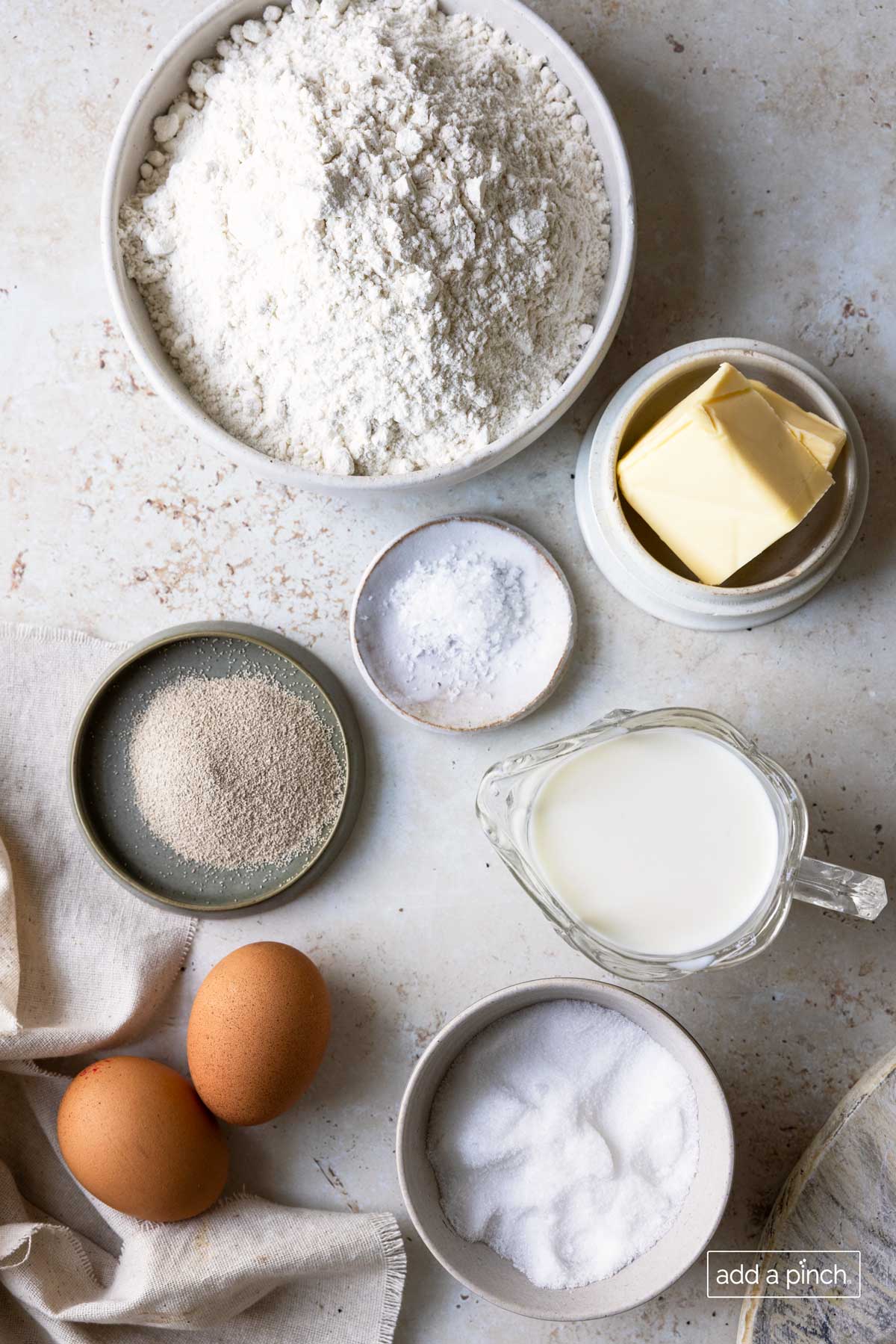 Marble countertop holds containers of ingredients for Cinnamon Roll recipe - flour, yeast, butter, cinnamon, sugar, eggs, salt, and milk.