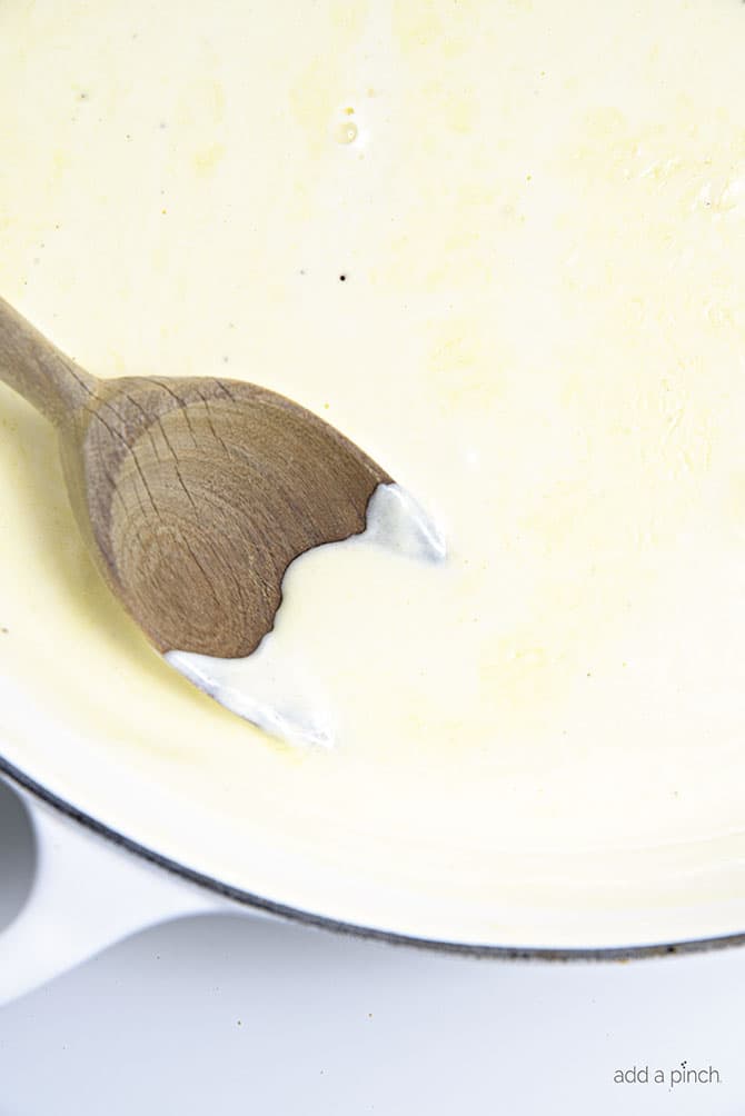 Alfredo sauce cooking and being stirred with a wooden spoon in a white cast iron skillet // addapinch.com