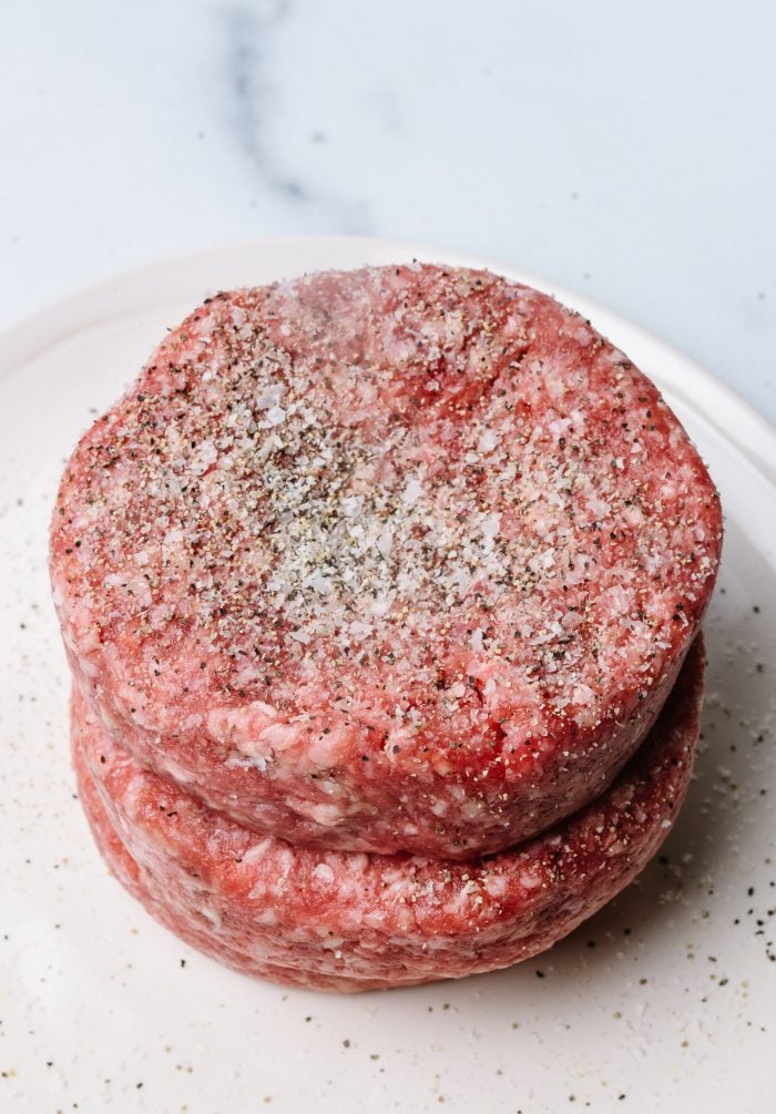 Ground chuck formed into three even thickness, 1/4 pound patties, seasoned with Stone House Seasoning, on white plate // addapinch.com