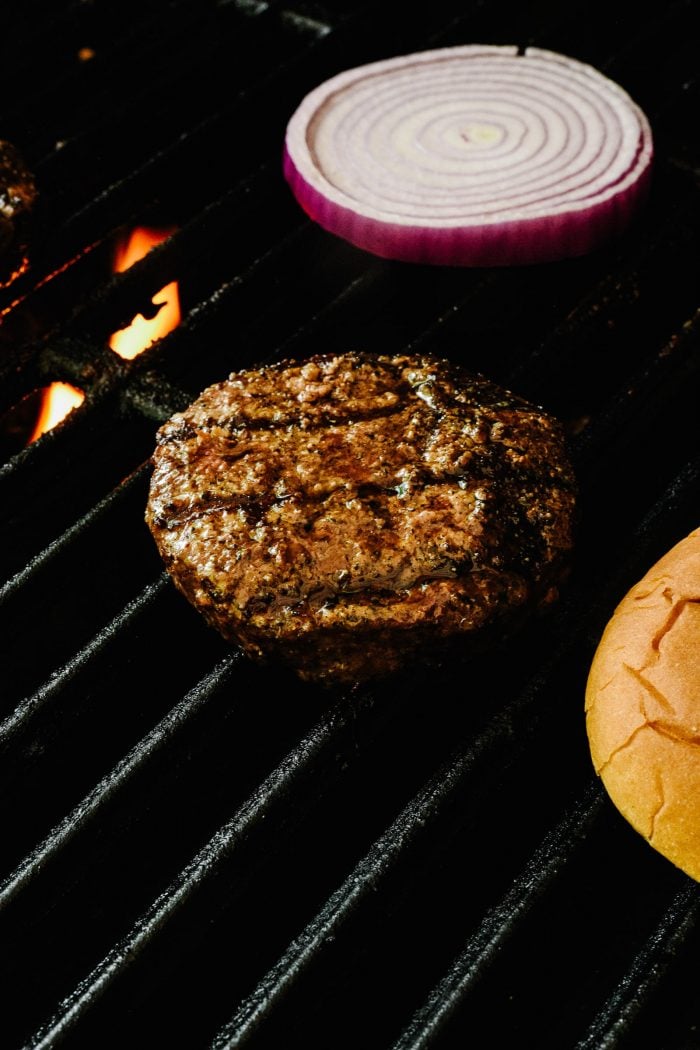 Grill grate with flame under it, with juicy grilled burger, onion slice and bun on grate.