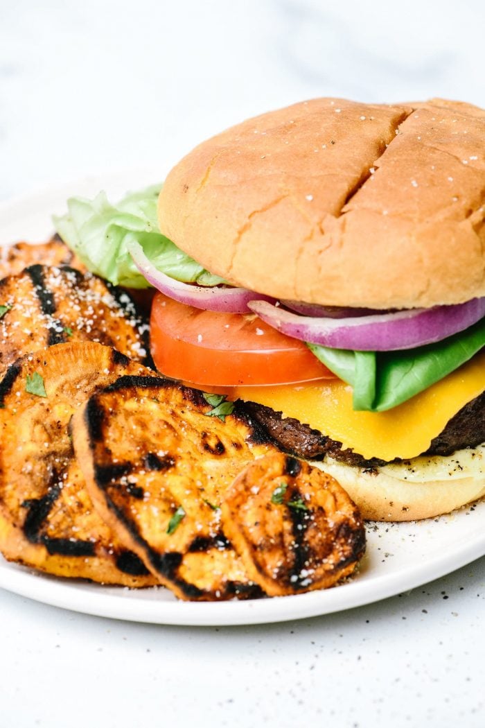 Hamburger on bun with cheese, sliced tomato, red onion, lettuce and side of grilled sweet potatoes on white plate.