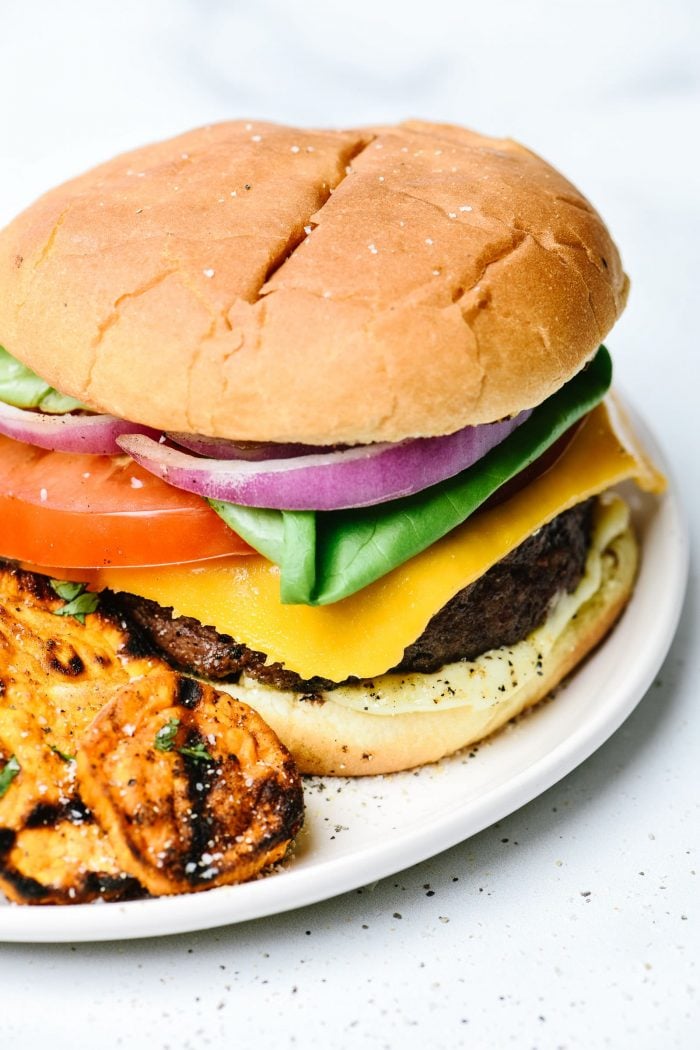 Hamburger on bun with all the toppings - onion, tomato, lettuce, cheese, mayo, served with grilled sweet potatoes on white plate.