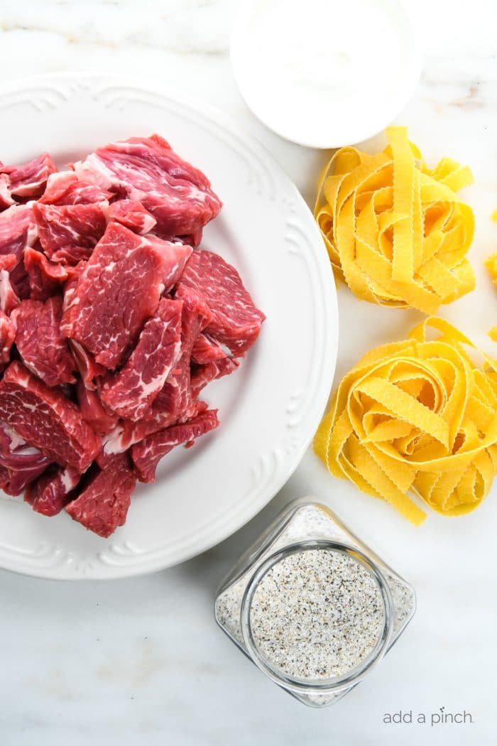 Photograph of sliced beef on a white plate with Stone House Seasoning in a jar, nests of egg noodles, and sour cream surrounding the plate on white marble.