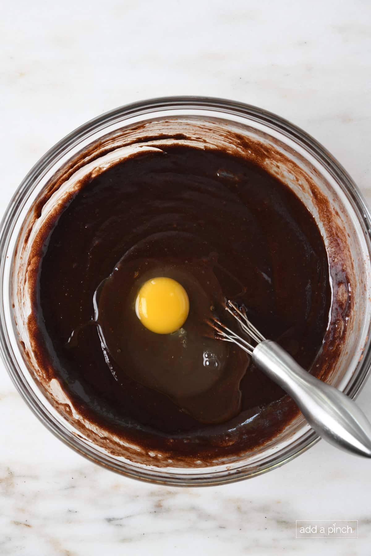 Glass mixing bowl with chocolate brownie batter with egg added ready to whisk into batter.