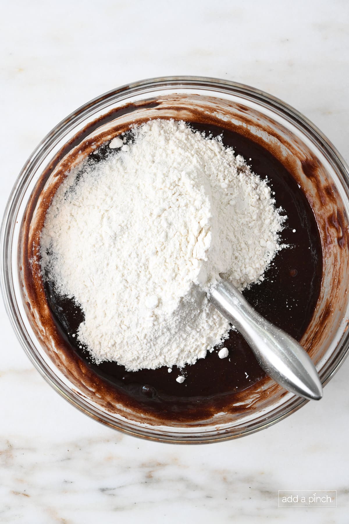 Marble countertop holds glass mixing bowl with chocolate brownie mixture with flour added ready to mix.