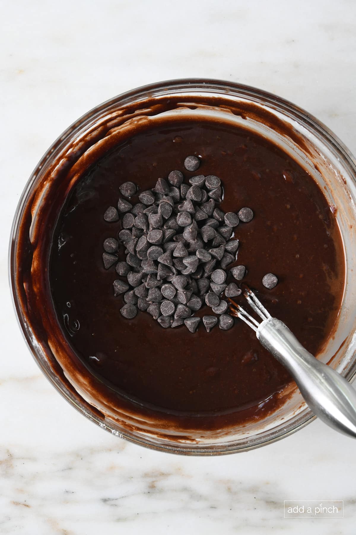 Glass bowl of brownie batter mixing in dark chocolate chips.