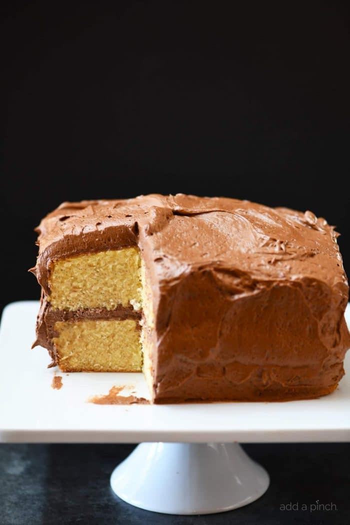 Vanilla cake frosted with chocolate buttercream has a large slice cut out of it as it sits on a white cake stand.