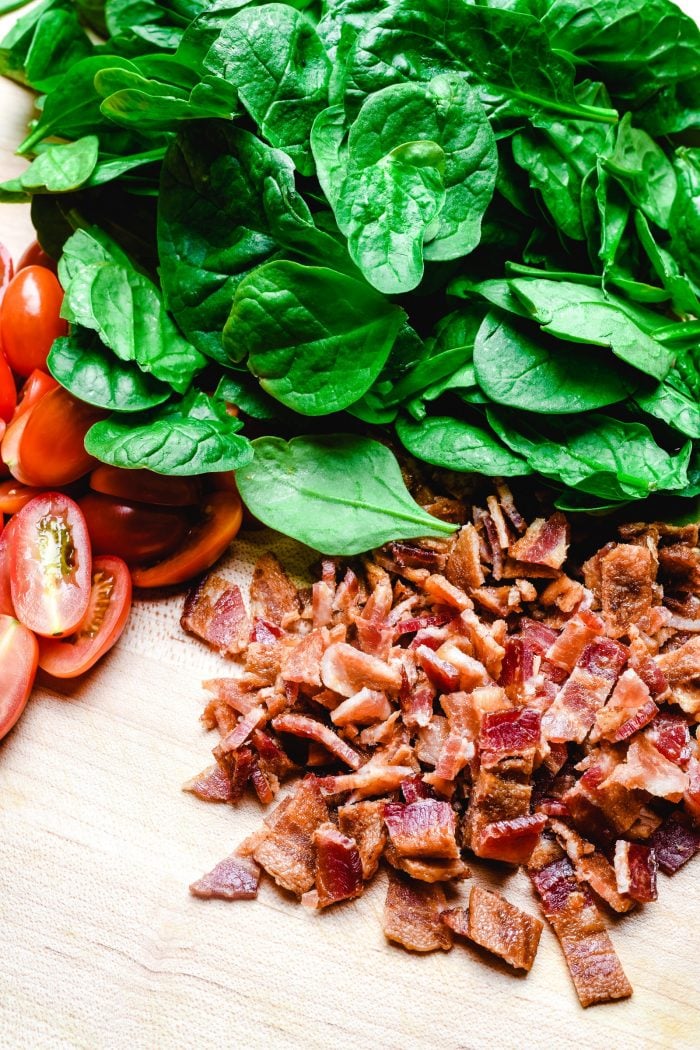Crisp pieces of bacon, spinach and sliced cherry tomatoes sit on a wooden cutting board.