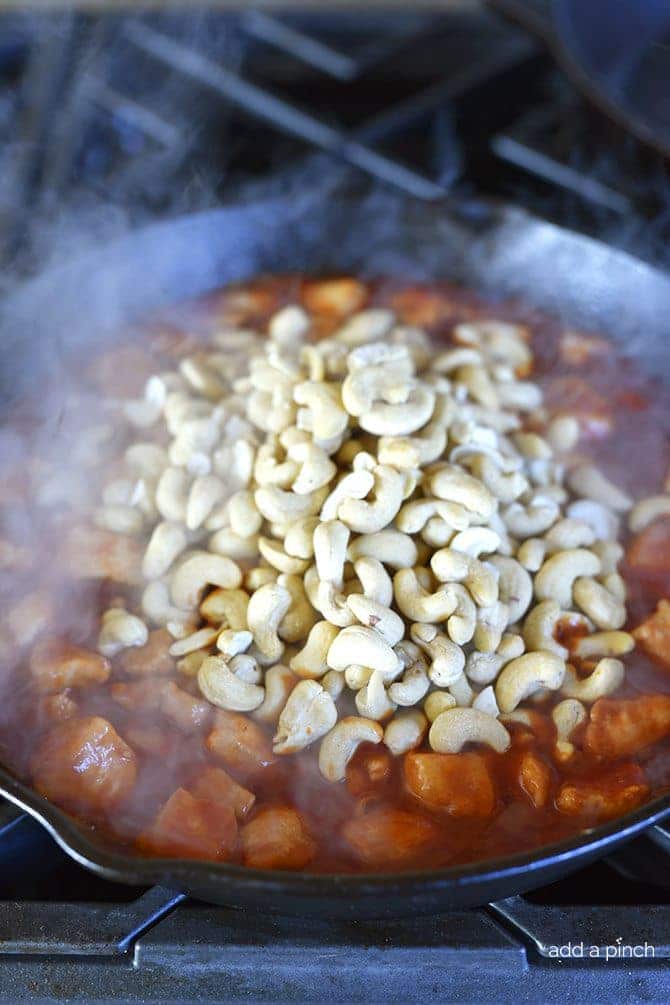 Raw cashews are added to skillet of cooking chicken in sauce.