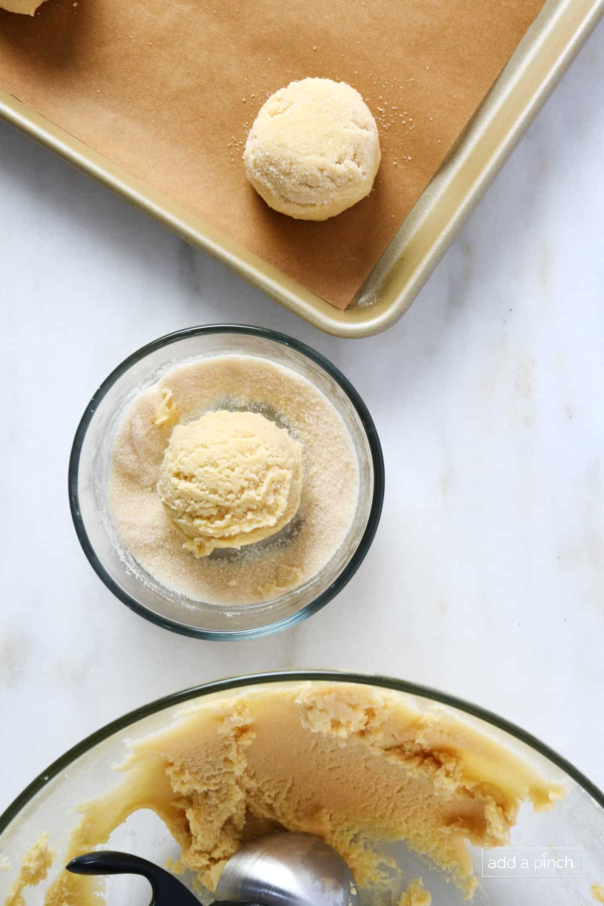 Scoop of chewy sugar cookie in a small glass bowl of sugar with larger bowl of dough to one side and baking sheet with more cookies on it.