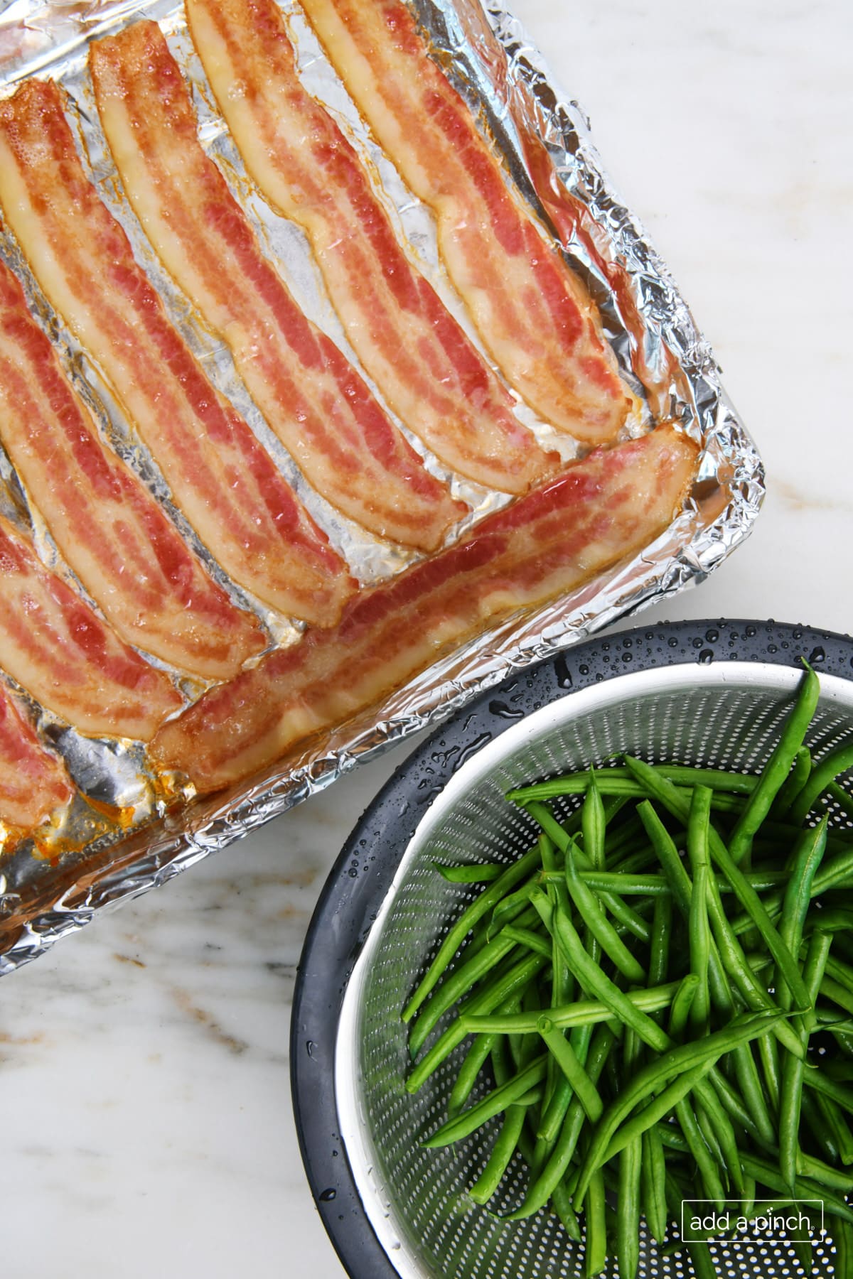 Bacon on a baking sheet and blanched green beans in a colander on a marble surface.