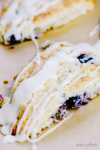 Scones filled with blueberries and drizzled with glaze are on a parchment lined sheet.