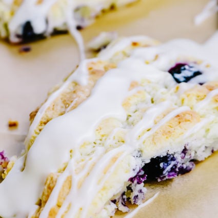 Scones filled with blueberries and drizzled with glaze are on a parchment lined sheet.