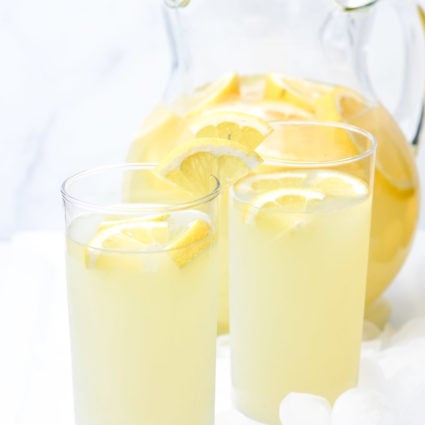 Photo of a clear pitcher of lemonade with lemon slices and 2 clear glasses of lemonade with lemon slices on a white background.