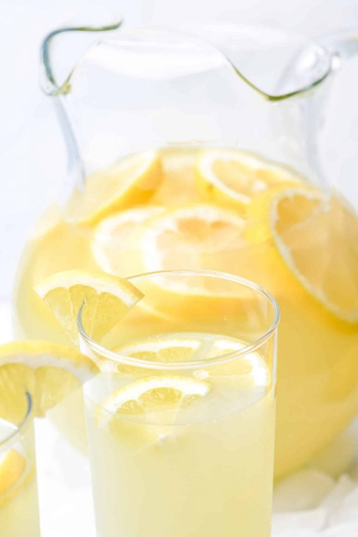 Pitcher of lemonade with fresh lemon slices sits beside two glasses filled with it and garnished with lemon slices.