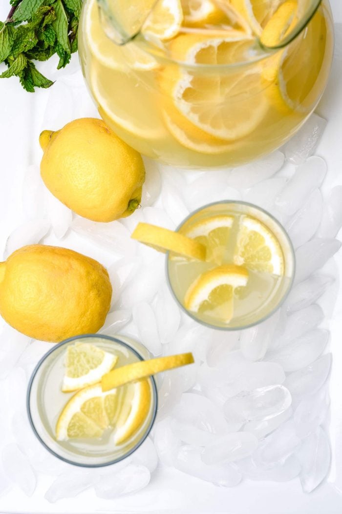 Overhead picture of a glass pitcher of lemonade, lemons, and glasses of lemonade with lemon slices. Mint garnish lies beside the pitcher.