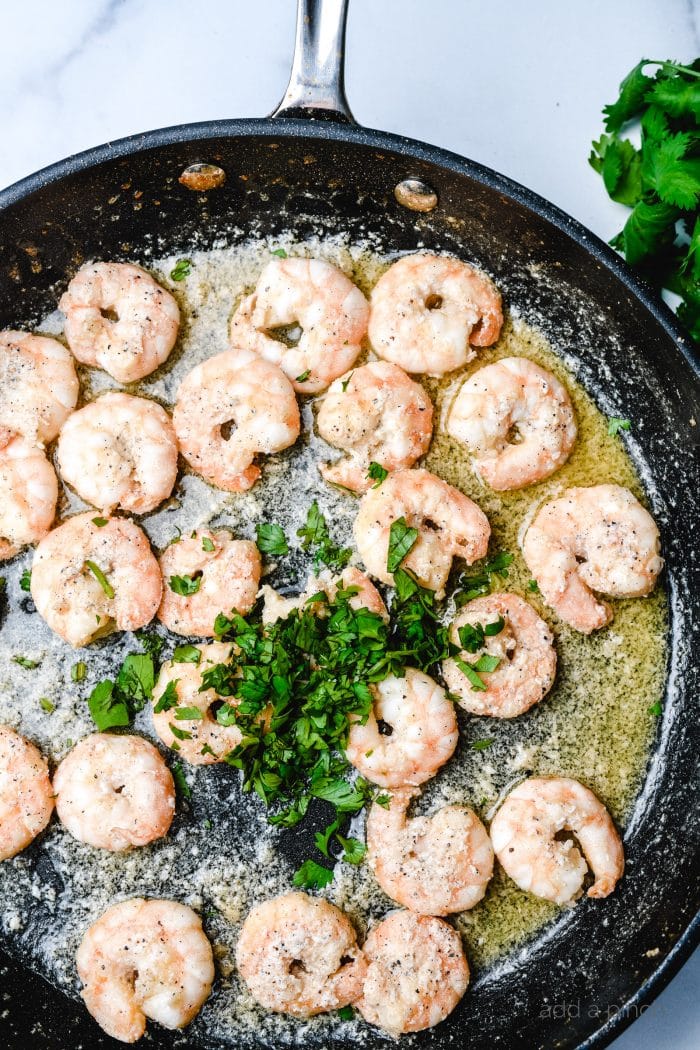 Skillet with melted butter and spices, cooking shrimp and topped with fresh parsley.