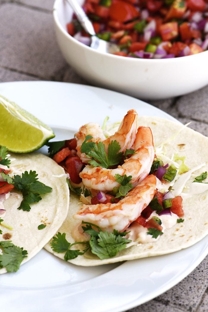 Shrimp tacos on a plate with a lime wedge, sitting beside a bowl of fresh pico de gallo.