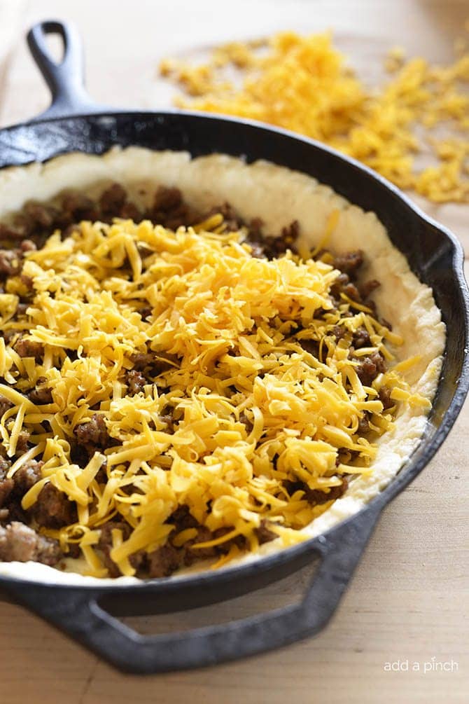 A cast iron skillet holds a biscuit crust filled with browned sausage and freshly grated cheddar cheese, while more grated cheese sits nearby on the wooden countertop.