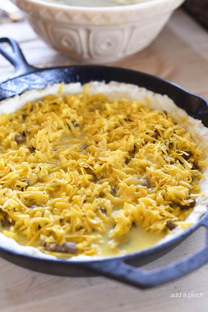 Shredded cheese tops egg mixture and browned sausage on a biscuit crust in an iron skillet as it is ready to go into the oven to bake.