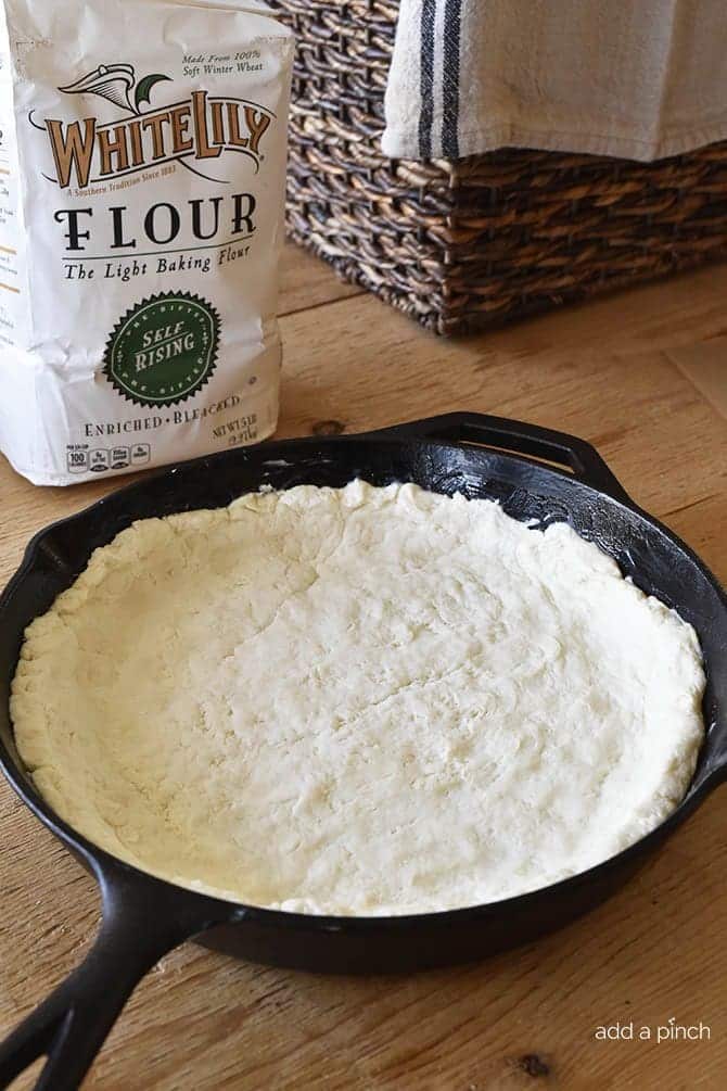 A large cast iron skillet holds cream biscuit dough to make a crust for quiche. A bag of self-rising flour sits close by the skillet and all are placed on a wooden board.