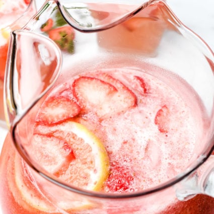 Glass pitcher is filled with ice, lemon and strawberry slices and strawberry lemonade, sits beside glasses filled with it too.