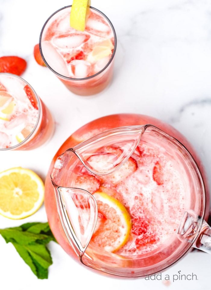 Top view of pitcher of garnished strawberry lemonade surrounded by lemon slices, glasses and mint sprigs.