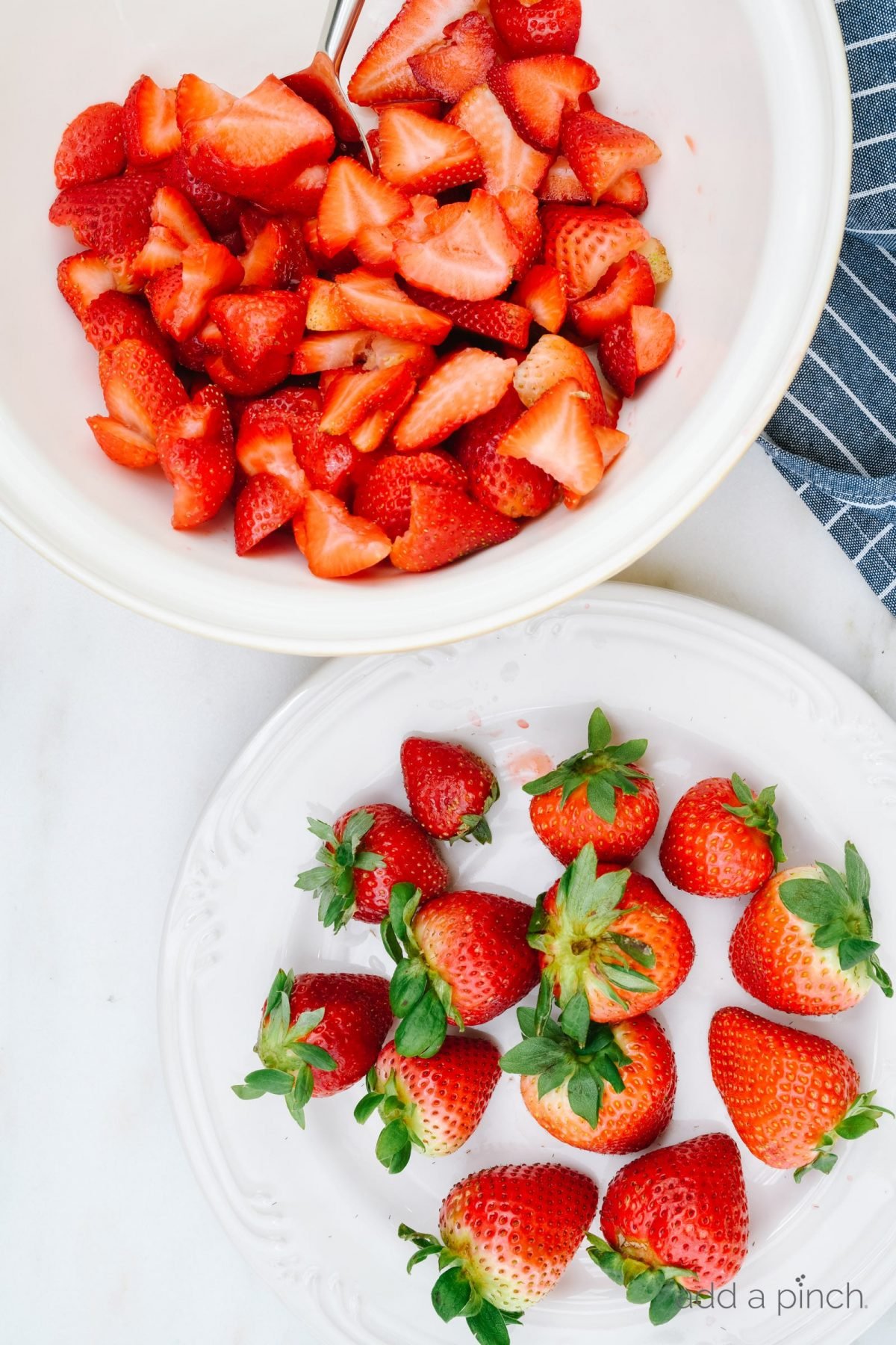 Photograph of strawberries on a white plate and sliced strawberries in a white bowl on a white marble background.