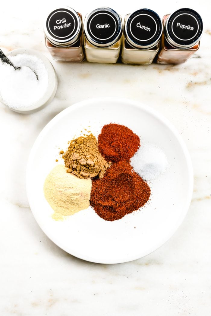 Plate holds four spices (chili powder, garlic powder, cumin, and paprika) and salt. There are labeled glass spice jars for each of these spices, as well as a salt cellar with a spoon behind the plate on this marble countertop.