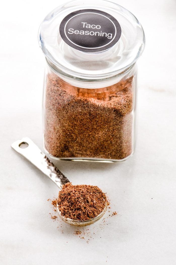 Square glass jar with lid labeled Taco Seasoning with a spoon holding the seasoning laid beside the jar.